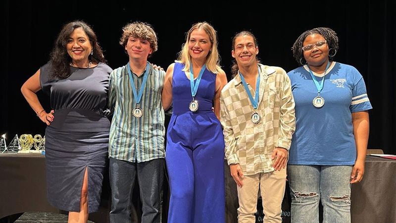Four students on stage smiling and wearing their medals around their necks, accompanied by a high school administrator
