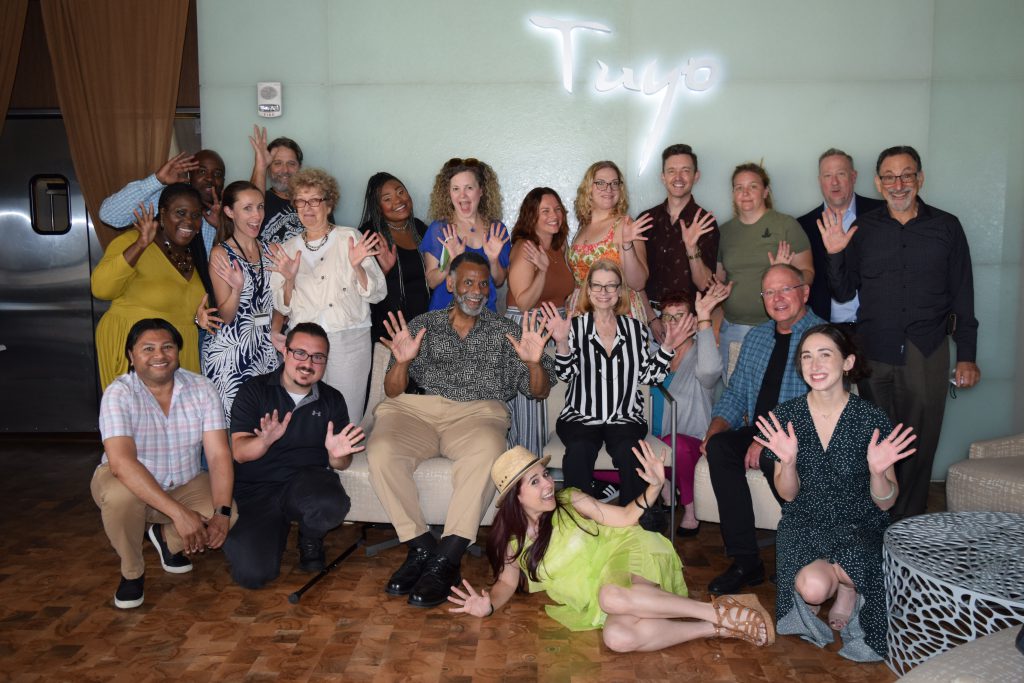 Large group of people, some sitting and some standing, smiling at the camera waving jazz hands.