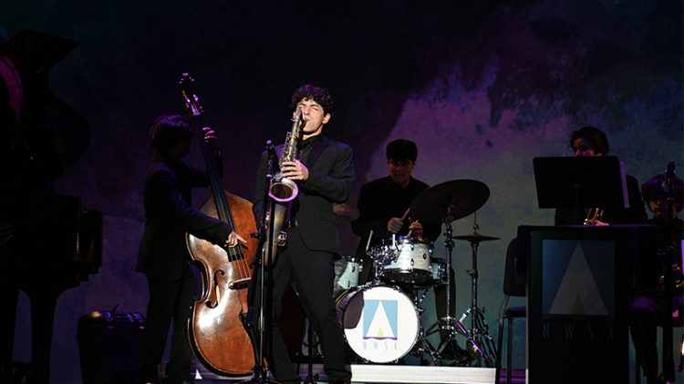 Student on stage playing the saxophone with silouette of other students playing instruments in the background