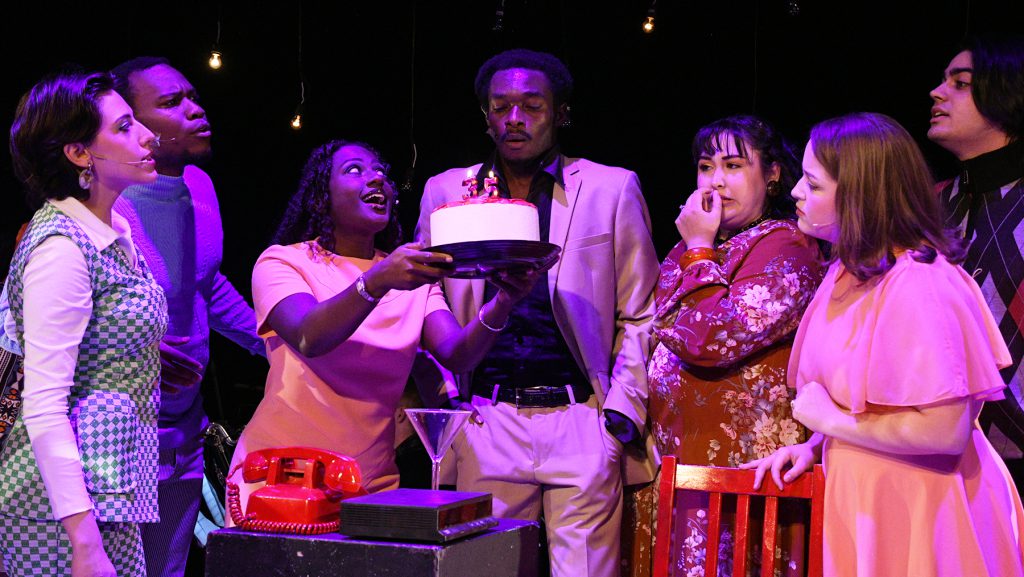 Seven students performing in Company. One holding a cake in front of another student.