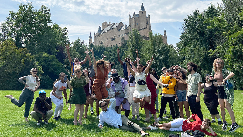 Large group of students jumping, standing or sitting on the grass as they pose in a park in front of a castle.