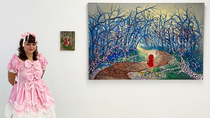 Student in pink outfit and pink head bow standing beside a small painting and a larger painting, both hanging on the wall.