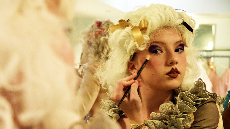 Student actor applying makeup in front of mirror in period costume and wig