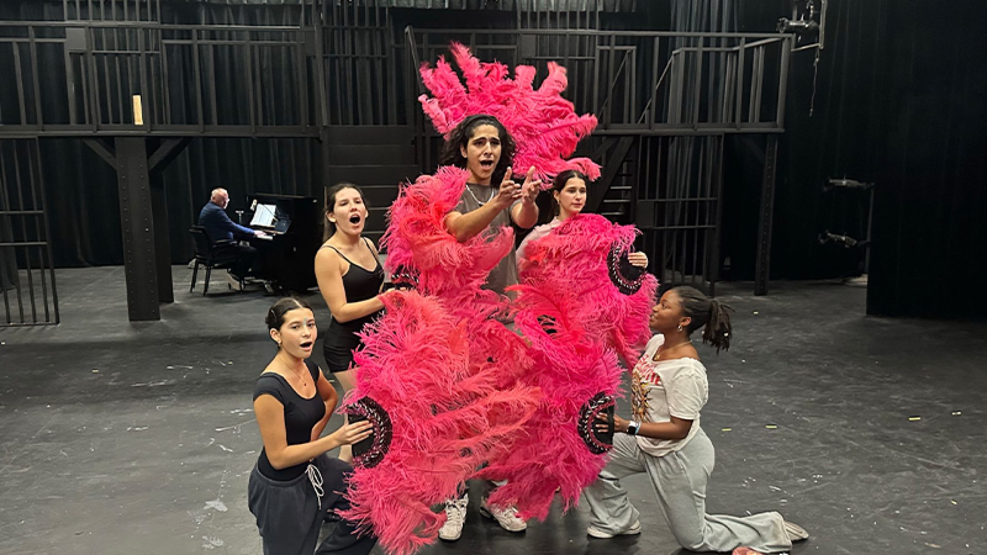 5 students rehearsing on stage with one sporting pink feathers. Person playing the piano in the background