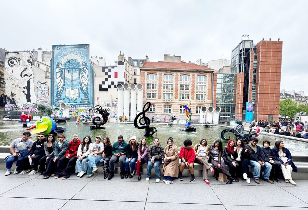 Group of students sitting in a line in front of a large fountain displaying black and multi-color sculptures.