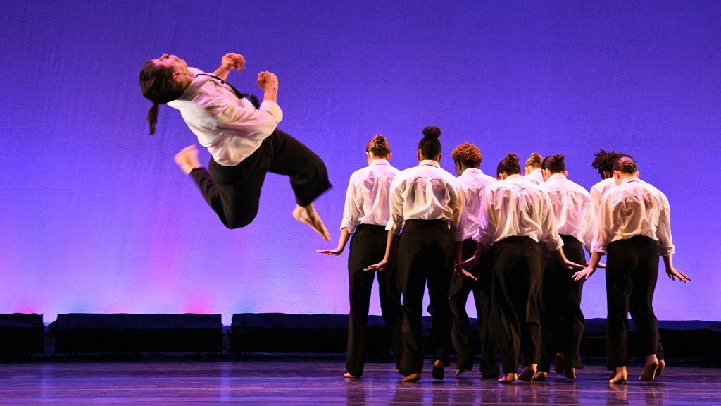 Nine dancers dancing on stage dressed in white and black with one dancer leaping and turning in the air.