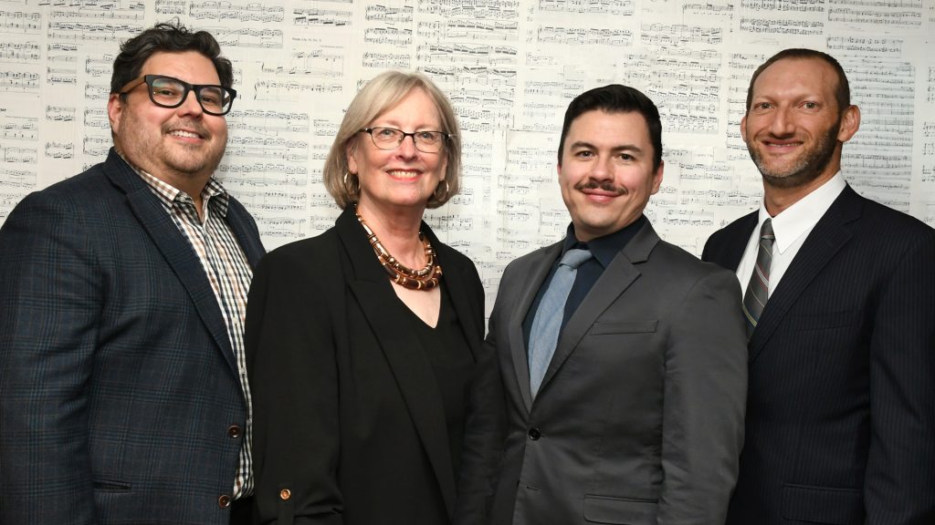 Four people smiling standing in front of a music sheet backdrop.