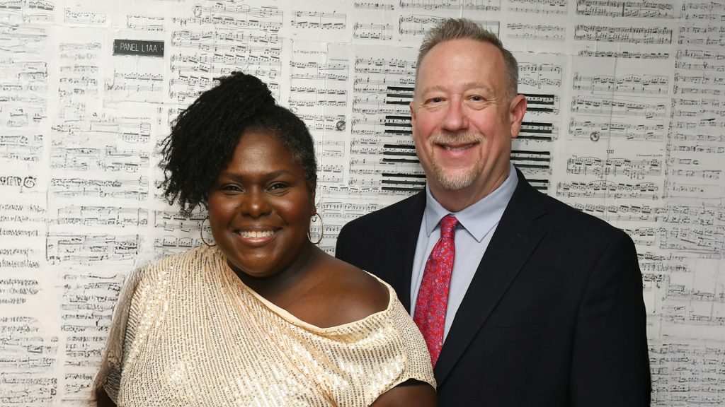 Two people smiling standing in front of a music sheet backdrop.