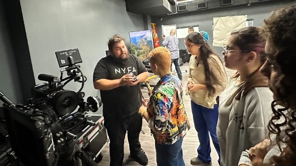 Seven people in a room with a video camera and a painting in the background.
