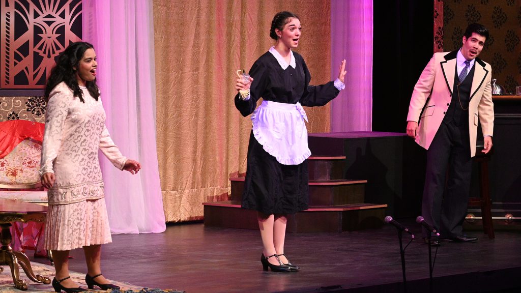 Three singers in 1920s costumes singing on stage.