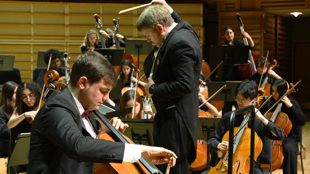 Student orchestra lead by director with baton at hand playing on stage with a cellist in the foreground.