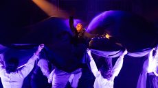 Actor in center surrounded by flowing skirt carried by other actors.