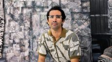 Person with glasses sitting in from of grey painting.