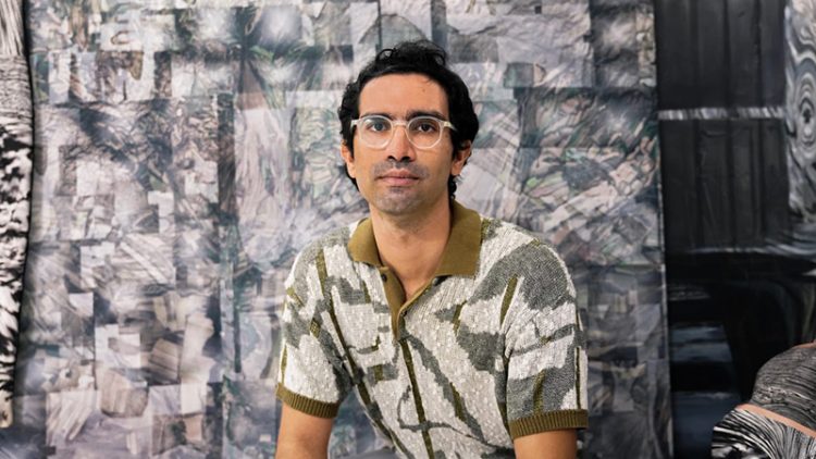 Person with glasses sitting in from of grey painting.